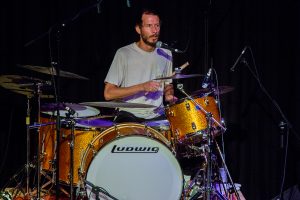 Mann mit lockigen Haaren spielt E-Gitarre und steht vor einem Mikrofon auf der Bühne.