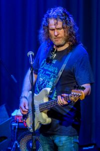 Mann mit lockigen Haaren spielt E-Gitarre und steht vor einem Mikrofon auf der Bühne.