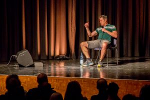 Ein Künstler steht auf der Bühne vor einem großen Publikum in einem Veranstaltungsraum.