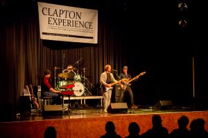 Band performing live on stage at the Clapton Experience tribute event with instruments and audience visible.