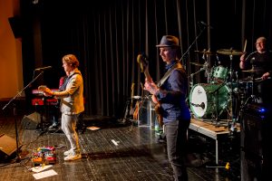 Drei Musiker auf einer Bühne, einer spielt Gitarre, ein anderer Bass, und der dritte sitzt am Schlagzeug.