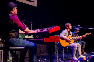 Drei Musiker auf der Bühne, einer spielt Keyboard, zwei andere Gitarre und Gesang.