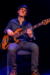 Mann mit Hut spielt Bassgitarre auf der Bühne während eines Live-Auftritts.