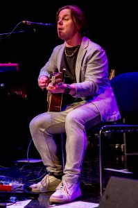 Ein Musiker sitzt auf einem Stuhl und spielt Akustikgitarre während eines Auftritts auf der Bühne.